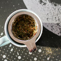 A cup of Cycle Goddess herbal tea by Lake Missoula Tea Company in a white mug with tea leaves and flowers on a wooden surface.