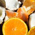 A close-up arrangement of fresh citrus fruit, including a large orange slice in the foreground, orange segments, coconut halves, and dried orange peel.