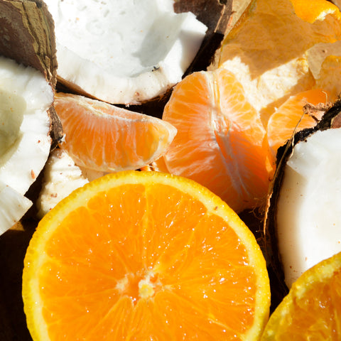 A close-up arrangement of fresh citrus fruit, including a large orange slice in the foreground, orange segments, coconut halves, and dried orange peel.