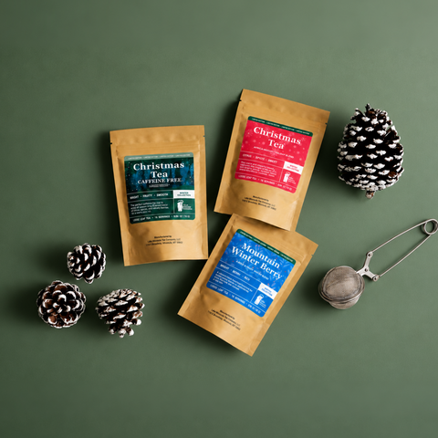 Three Lake Missoula Tea Company winter collection loose-leaf tea pouches—Christmas Tea, Christmas Tea Caffeine Free, and Mountain Winter Berry—arranged on a muted evergreen background with a stainless steel tea infuser and snow-dusted pinecones.