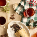 A cozy flat-lay scene featuring tea products, including loose leaf tea in kraft paper pouches and cups of brewed tea, arranged with star anise, greenery, a knit blanket, and holiday cookie cutters on a beige fabric surface.