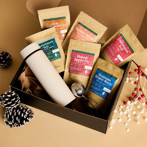 Holiday Tea Bundle gift box with seasonal tea pouches, a white tumbler and a tea infuser, surrounded by pinecones