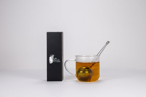 Glass mug with tea and a tea bag, next to a black box with branding on a white background