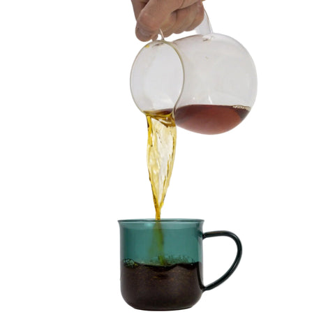 Glass teapot pouring loose leaf tea into a Viva Minima mug on a white background