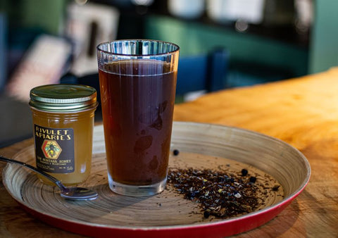 Chilled Gin & Cream Rooibos tea served in a glass, with loose tea leaves and honey spoon on a wooden surface.