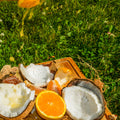 A wooden cutting board outdoors on green grass, holding halved coconuts, a sliced orange, and a stream of orange juice splashing down from above.