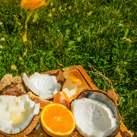 A wooden cutting board outdoors on green grass, holding halved coconuts, a sliced orange, and a stream of orange juice splashing down from above.