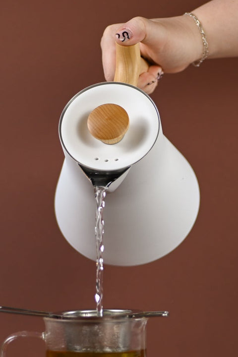 White Fellow Corvo electric water heater kettle pouring hot water into glass tea brewer, showcasing sleek teaware design.