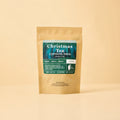 A stand-up kraft paper pouch of Christmas Tea Caffeine-Free from Lake Missoula Tea Company, featuring a dark teal festive label with white text and a winter forest design, set against a solid pale peach background.